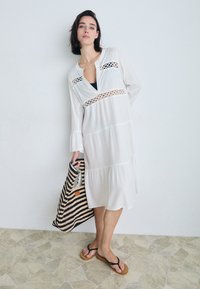 Vestido branco de mangas longas com detalhes em renda, decote em V e mangas com babados. Combinado com uma bolsa de mão às riscas e chinelos.