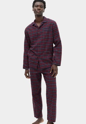 Man standing barefoot wearing red and navy blue plaid long-sleeve button-up pajama top and matching pants on a plain background.