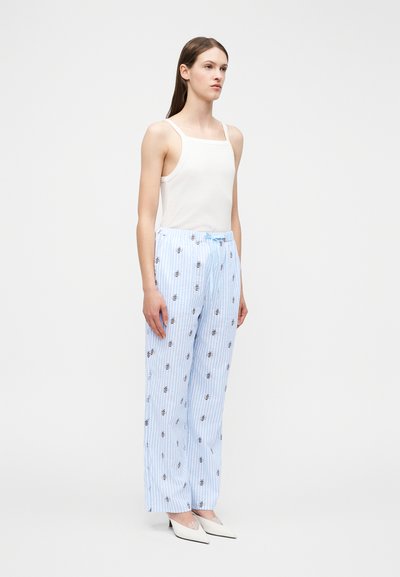 Kvinde iført en hvid ærmeløs top, lyseblå bukser med vide ben og bi-mønster samt hvide spidse hæle, stående mod en ensfarvet baggrund.