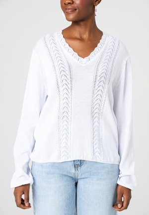 Kobieta w białym swetrze z dekoltem w serek, ozdobionym koronką i ażurowym wzorem, w połączeniu z jasnoniebieskimi dżinsami, na jednolitym tle.