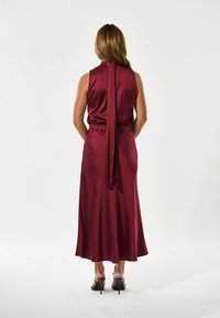 Burgunder satin maxikjole med høj halsudskæring, åben ryg og bånddetalje. Har en elastisk talje og en flowy skirt. Sorte hæle båret.