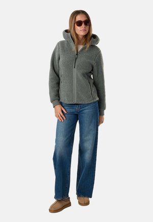 Kvinde iført solbriller, grå fleecejakke med lynlås og hætte, blå jeans og brune slip-on sko, stående med den ene hånd på hoften.