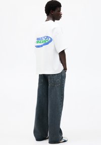 White oversized t-shirt with "CREATIVE MINDSET" graphic in blue and green. Worn with wide-leg dark denim jeans, side pockets visible.