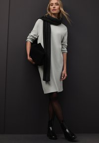 Grijze sweatshirtjurk met lange mouwen, gecombineerd met een zwarte sjaal en clutch. Model draagt zwarte enkelharen met een glanzende afwerking.