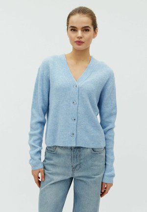 Mladi žena u svijetloplavom kardiganu na kopčanje i plavim trapericama, stoji ispred jednobojne svijetle pozadine.