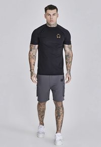 Czarny sportowy t-shirt z złotym logo na piersi, połączony z szarymi szortami cargo z kieszeniami. Materiały wyglądają na gładkie i oddychające.