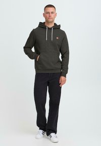 Man wearing dark gray hoodie, black jeans, and white sneakers with black stripes, standing with one hand in pocket on white background.