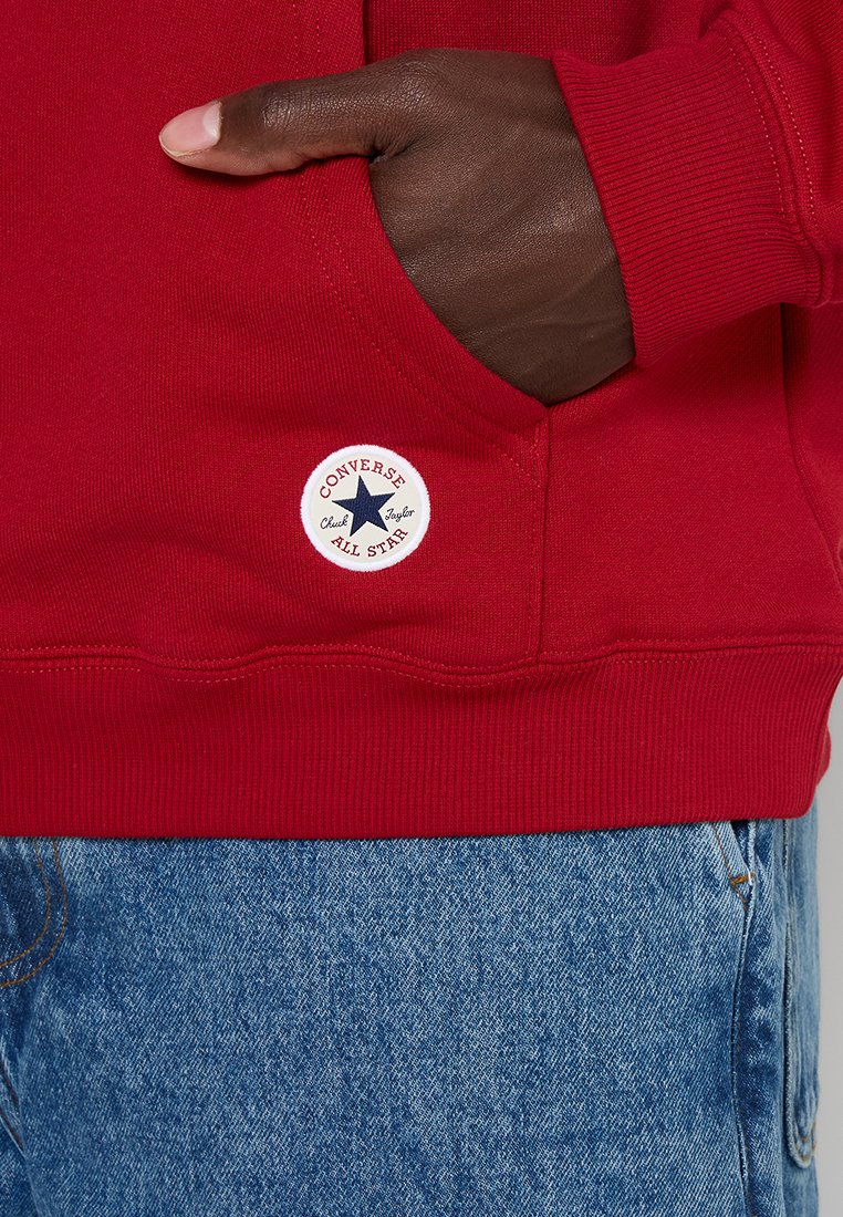 Czerwona bluza z ribbowymi mankietami i dołem, z okrągłą białą łatą z niebieską gwiazdą i napisem "Converse Chuck Taylor". Noszona z niebieskimi dżinsami.