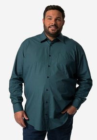 Camicia a maniche lunghe di colore verde scuro in tessuto morbido, con taschino sul petto e bottoni neri, abbinata a jeans blu.