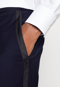 Marineblauwe broek met zwarte satijnen zijstreep; slank ontwerp; stof lijkt glad met een iets gestructureerde afwerking; hand in zak.