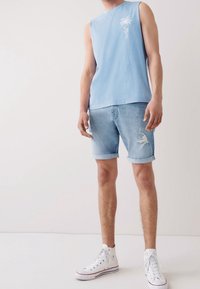 Chemise sans manches bleu clair avec broderie de palmiers, associée à un short en denim bleu clair déchiré, le tout complété par des baskets montantes blanches.