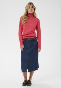 Pull à col roulé rouge, jupe en jean avec des rayures verticales, fente jusqu'aux genoux. Ceinture marron et chaussures avec détails. Fond neutre.