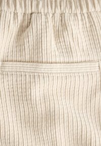 Tela de sarga color crema con textura de ribete vertical. Cintura elástica y un detalle de costura horizontal encima de un bolsillo.