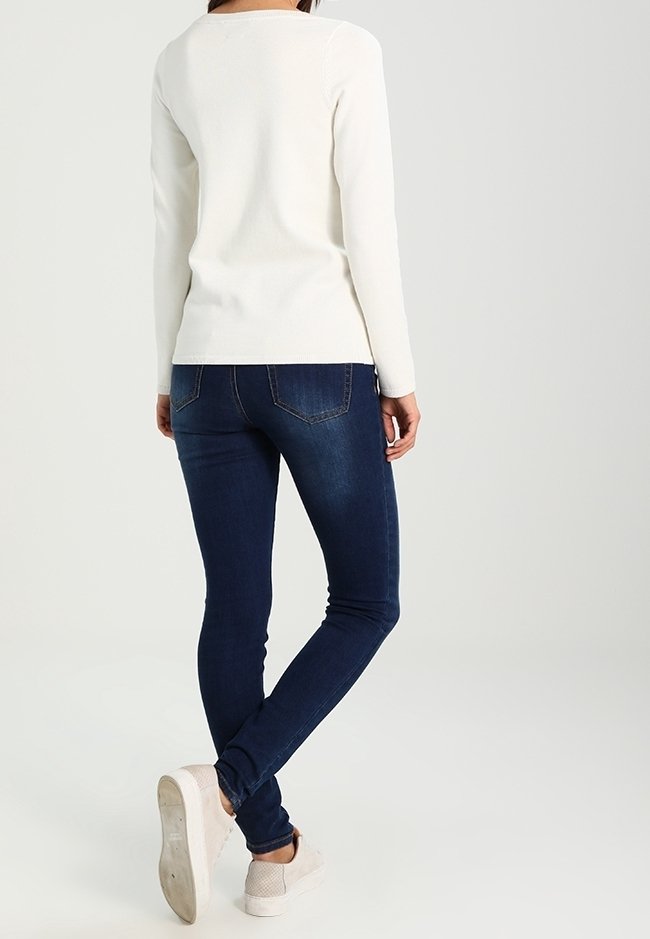 Femme portant un haut blanc à manches longues, un jean skinny bleu foncé et des baskets beige clair, debout avec une jambe croisée derrière l'autre.