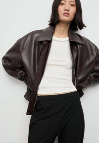 Brązowa skórzana kurtka pilotka z dużym kołnierzem, zamkiem błyskawicznym i ściągaczem, w połączeniu z białą dopasowaną bluzką i czarnymi spodniami.