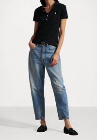 Svart polo med korta ärmar och knäppning framtill, kombinerad med ljusblå, löst sittande jeans. Svarta slip-on skor fulländar stilen.