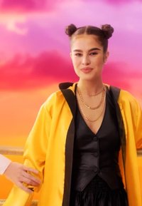 Ung kvinne med to toppers som har lag med gullkjeder, svart vest og lys gul jakke foran en livlig rosa og oransje himmelbakgrunn.