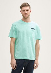 Camiseta de algodón verde menta clara con escote redondo, mangas cortas y un bolsillo en el pecho que presenta una etiqueta azul marino. Tejido texturizado.
