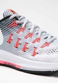 Gros plan d'une chaussure de course Nike Zoom gris clair avec des œillets de lacets orange, des lacets blancs et des détails noirs et roses sur la semelle.