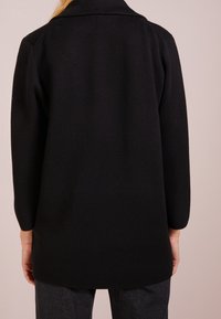 Svart långärmad kofta med krage, texturerad tyg, rak passform och medellång design. Bärs över mörka jeans.