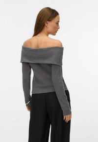 Kobieta z długimi włosami ubrana w szary sweter z odkrytymi ramionami i czarne spodnie, stojąca lekko bokiem na białym tle.