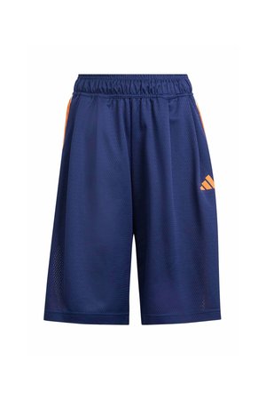 Adolescente con ropa deportiva azul de Adidas con pantalones cortos y camiseta, sosteniendo una gorra naranja en una mano, zapatillas blancas y calcetines a rayas.