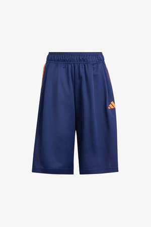 Adolescente con ropa deportiva azul de Adidas con pantalones cortos y camiseta, sosteniendo una gorra naranja en una mano, zapatillas blancas y calcetines a rayas.