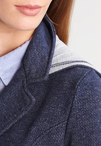 Marinblå jacka med en texturerad yta, prydd med en grå huva och kontrasterande sömmar. Närbild på kragen och skjortan under.