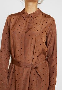 Braunes langärmeliges Kleid mit Kragen, ausgestattet mit einer Knopfleiste und schwarzen Punkten, an der Taille mit einem passenden Stoffgürtel gerafft.