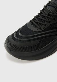 Black athletic shoe with mesh and rubber materials. Features curved design, textured accents, and a padded collar. Narrow laces and rubber sole.