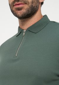 Gros plan d'un homme barbu portant un polo vert texturé avec une demi-fermeture éclair argentée au col.