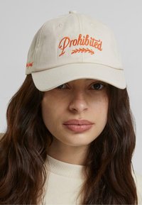 Beige baseball sapka, melyen narancssárgával hímezve az "Ingyenes" felirat található, ívelt karimával és állítható pánttal a hátsó részen.