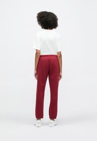 Άτομο με σγουρά μαλλιά, γυρισμένο με την πλάτη, φοράει λευκό κοντομάνικο μπλουζάκι, μπορντό φόρμα, και λευκά αθλητικά παπούτσια σε απλό φόντο.