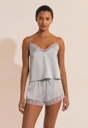 Frau trägt ein hellgraues Satincamisole- und Shorts-Set mit Spitzenbesatz, steht vor einem beigen Hintergrund und blickt nach vorne.