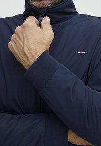 Tumši zilas krāsas pūka jaka ar augstu apkakli, ar rāvējslēdzēju un ribinātām piedurknēm. Krūts daļā ir neliels sarkans, balts un zils logos.