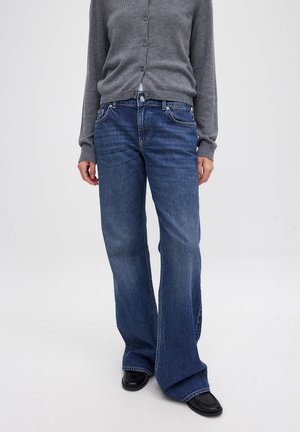 Flared blue denim jeans with a mid-rise, front pockets, and subtle stitching details. Paired with a gray cardigan and black shoes.