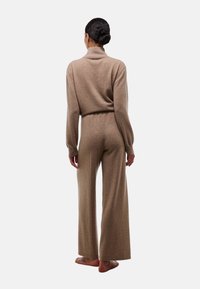 Une combinaison en tricot beige avec un col montant, des manches longues et un pantalon large, fabriquée en tissu doux et texturé. Vue de dos affichée.