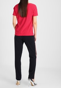 Red short-sleeve t-shirt paired with black pants featuring side stripes. The model wears silver high-heeled shoes. Background is neutral.