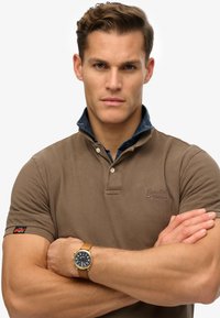 Polo shirt marrone con colletto in denim, maniche corte e logo sul petto. Il modello indossa un orologio dorato con quadrante blu e cinturino in pelle.