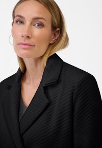 Blazer nero con un motivo a spina di pesce texturizzato, colletto affilato e spalle strutturate; indossato sopra un top grigio.
