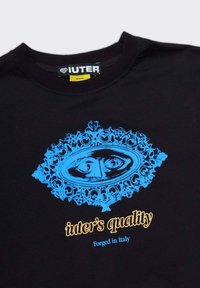 T-shirt nera in cotone con una grande grafica blu di un occhio all'interno di una cornice ornata, con il testo "qualità iuter" e "Forgiato in Italia" in giallo.