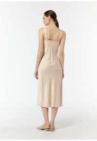 Beżowa satynowa sukienka midi z kwadratowym dekoltem, cienkimi ramiączkami i gładką teksturą. Noszona z minimalistycznymi beżowymi szpilkami.