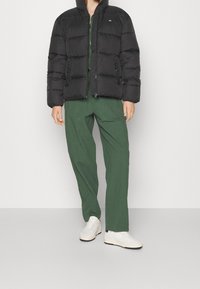 Svart pufferjacka med quiltat mönster och hög krage, matchad med gröna strukturerade vida byxor och vita sneakers.