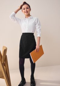 Hvid skjorte med knapper, sort pencil skirt, sorte strømpebukser og sorte loafers; holder en orange mappe, står nær en træstol.