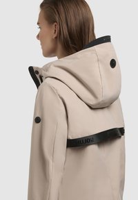 Veste imperméable beige clair avec capuche, présentant des accents noirs et une sangle de marque, texture lisse et détail boutonné sur la manche.