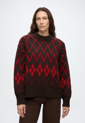 Femme aux longs cheveux bruns portant un pull marron avec des motifs en losanges rouges et zigzag, assorti à un pantalon marron, debout devant un fond uni.