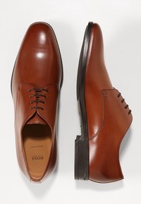 Chaussures Oxford en cuir marron à lacets avec semelles noires, une chaussure montrée de vue de dessus et l'autre de vue de côté.