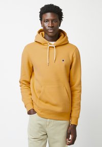 Felpa gialla in cotone con tasca a marsupio, cordino bianco e un piccolo emblema di un albero sul petto. Il modello indossa pantaloni chiari.