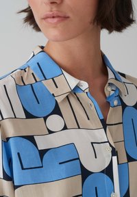Camicia button-up con motivi astratti in blu, beige e nero. Tessuto liscio con colletto classico e patta dei bottoni in vista.