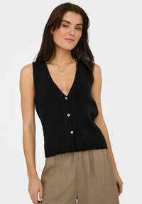 Chaleco de punto negro con escote en forma de V, que presenta un cierre de botones y un acabado texturizado. Llevado sobre pantalones beiges de corte suelto.